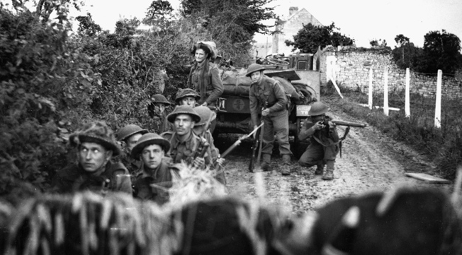With wary expressions, British infantrymen of the 130th Brigade, 43rd (Wessex) Division take cover in the shelter of a hedgerow during a mortar barrage, July 10, 1944. The 43rd Division was heavily engaged in the battle for Hill 112.